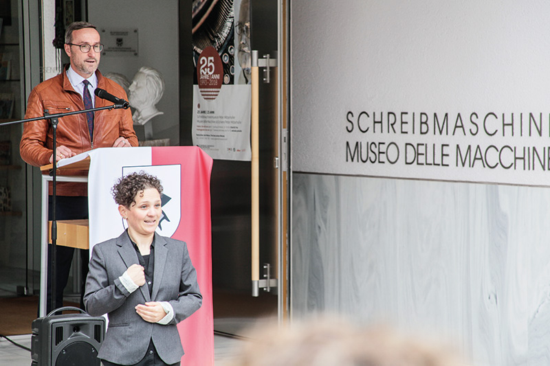 Gemeindereferent Ulrich Schweitzer begrüßt die Vertreter der Genossenschaft Independent L, die Vertreter der Stiftung Südtiroler Sparkasse, die Koordinatorin von Interreg ITAT Sabine Lamprecht, die Vertreter vom Palais Mamming in Meran. In Gebärdensprache übersetzt hat Julia Gamper.