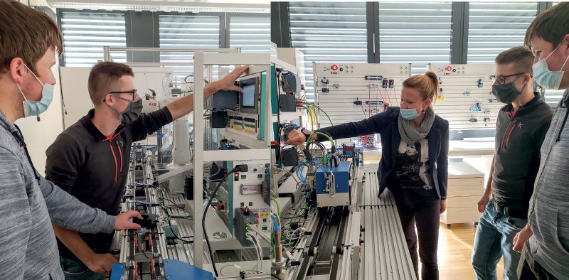 Daniel Gerstgrasser und Marian Mair bei der Eingabe des Auftrages; rechts: Erklärung der Qualitätssicherung mithilfe der Kamera durch Prof. Martina Trenkwalder. Im Hintergrund sieht man die Pneumatik- und Elektropneumatikstationen im Mechatroniklabor.