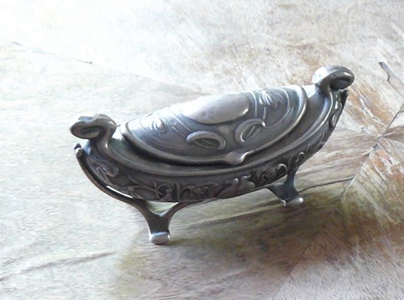 Silberne Schmuckdose auf Rauchertischchen: Der Jugendstil liebte aus kostbaren Hölzern geschnittene Furniere. Sie kamen aus aller Welt. Und so denke ich mit der „Silbergondel“ zugleich an die Maria Silbernagl, deren Grabstein sich lange an der Südseite der Pfarrkirche von Schlanders befand: Auffallend der Blumenschmuck mit der Lieblingsblume des Jugendstiles, der Schwertlilie. Sie ist die Verkörperung des Regenbogens, im Mittelalter eine Heilpflanze, daher ein Marien-Symbol. Diese Iris wählte man zum Wappen der Region Brüssel, weil sich in der europäischen Hauptstadt besonders viel Jugendstil befindet. Der griechische Name Iris bedeutet Regenbogen und ist Sinnbild für die Versöhnung zwischen Gott und den Menschen.