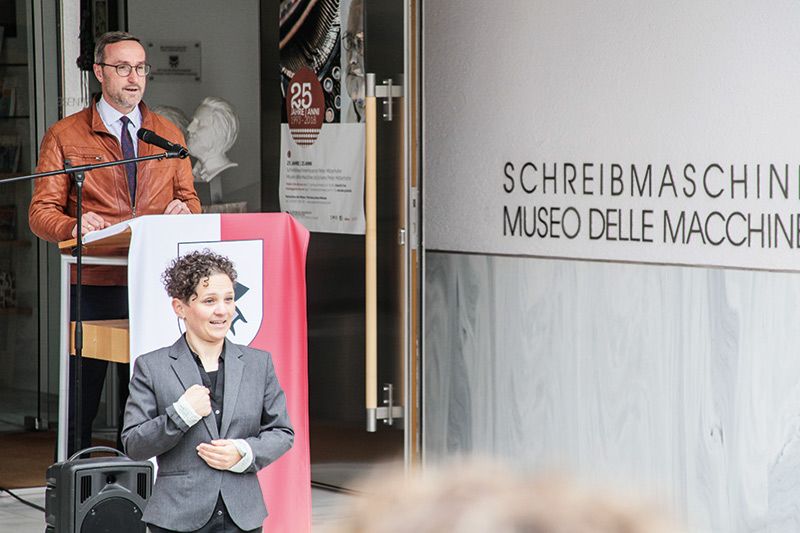 Gemeindereferent Ulrich Schweitzer begrüßt die Vertreter der Genossenschaft Independent L, die Vertreter der Stiftung Südtiroler Sparkasse, die Koordinatorin von Interreg ITAT Sabine Lamprecht, die Vertreter vom Palais Mamming in Meran. In Gebärdensprache übersetzt hat Julia Gamper.